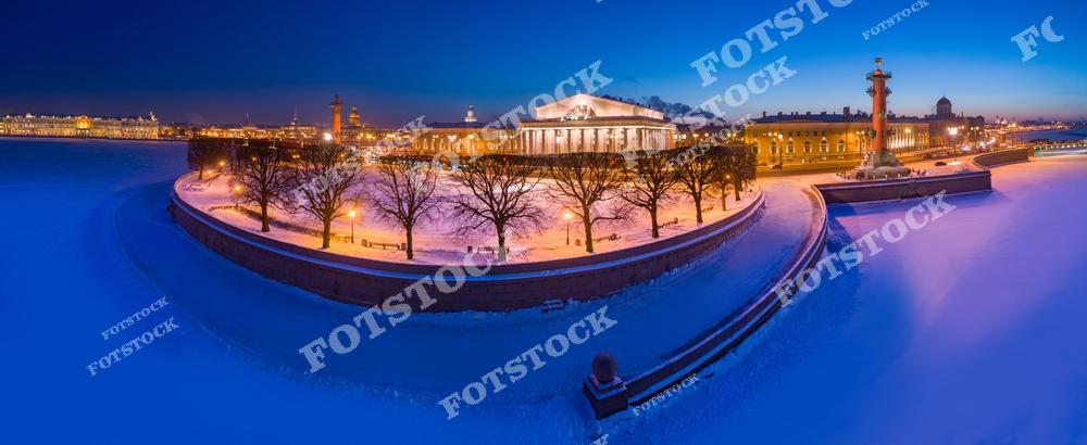 На фотографии запечатлена Стрелка Васильевского острова, окружённая заснеженной Невой. Яркие огни Ростральных колонн и архитектура здания Биржи создают контраст с холодной синевой ночного снега. Вид подчёркивает величественность и спокойствие зимнего Петербурга.